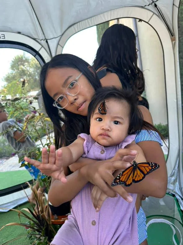 An older sister cradling a baby as monarch butterflies perch on them